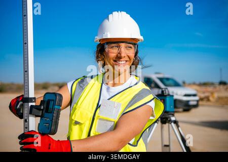Smiling female construction worker wearing a white helmet and safety vest, holding survey equipment on an outdoor job site, highlighting workplace saf Stock Photo