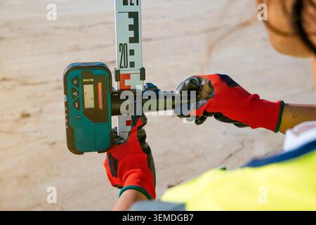 Female construction worker wearing safety gloves and vest uses a laser level with measuring rod on a building site, ensuring accurate alignment and sa Stock Photo