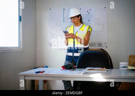Female construction worker wearing a safety helmet and reflective vest checks a smartphone in a site office, with blueprints on the wall and desk for Stock Photo