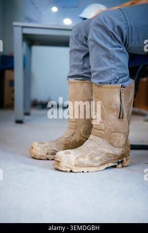 Close-up of muddy safety boots worn by a construction worker on an indoor job site, showing dirt, wear and protective footwear for building, repair an Stock Photo