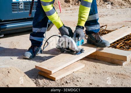 Close-up of a construction worker in high-visibility clothing cutting a wooden plank with a circular saw on a building site, with dust, tools, and mat Stock Photo