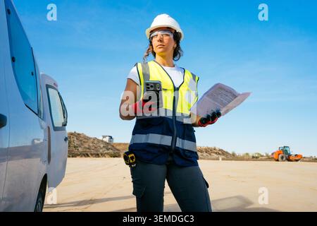 Confident female construction worker in hard hat and safety vest checks a smartphone while holding blueprints at an outdoor job site, with van and equ Stock Photo
