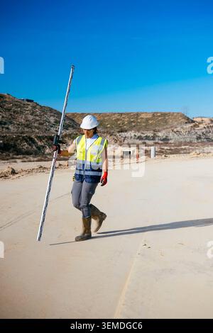 Female construction worker in hard hat and safety vest walks across a roadwork site holding a leveling rod, checking ground elevation under a clear bl Stock Photo