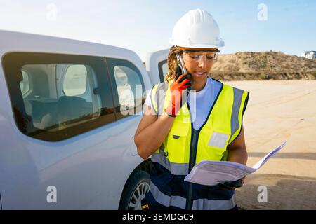 Female construction worker in hard hat and safety vest uses a smartphone beside a work truck, reviewing paperwork on an active outdoor construction si Stock Photo
