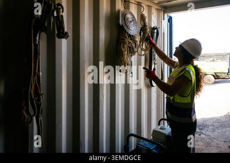A female construction worker in hard hat and high-visibility vest organizes ropes and safety equipment inside a site container, preparing tools for da Stock Photo