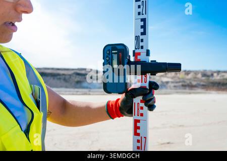 Female construction worker using a laser level and measuring rod to check elevation on a building site, wearing safety vest and gloves under clear blu Stock Photo