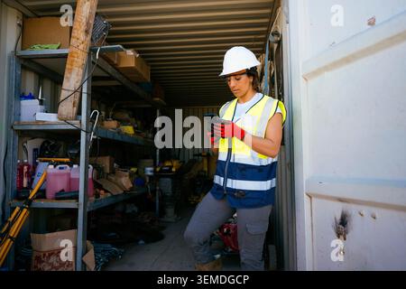 A female construction worker in hard hat and reflective vest stands in a storage container, checking a smartphone among tools and supplies at an activ Stock Photo