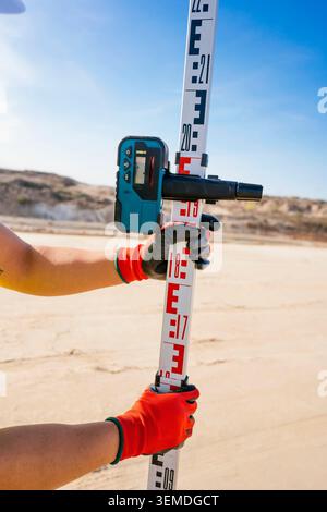 Close-up of a construction worker wearing safety gloves using a laser distance meter with a survey leveling staff on a sunny site, showing accurate me Stock Photo