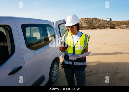 Female construction worker in hard hat and safety vest checks a smartphone while holding plans beside a work van at a sandy jobsite, managing tasks an Stock Photo