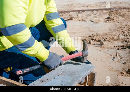 Close-up of a construction worker in high-visibility jacket using a hammer on wooden formwork at an outdoor building site, focused on hands, tools, an Stock Photo