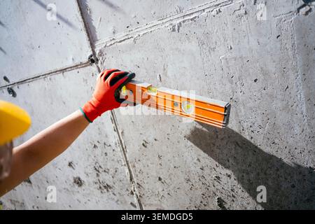 Close-up of a construction worker in safety gloves and helmet using an orange spirit level to check alignment on a raw concrete wall at a building sit Stock Photo