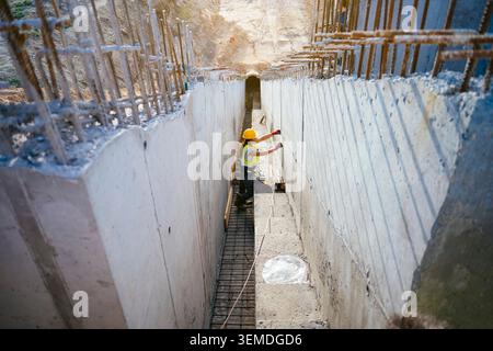 A woman construction worker in hard hat and safety vest measures and inspects a concrete trench with rebar, highlighting engineering work, safety, and Stock Photo