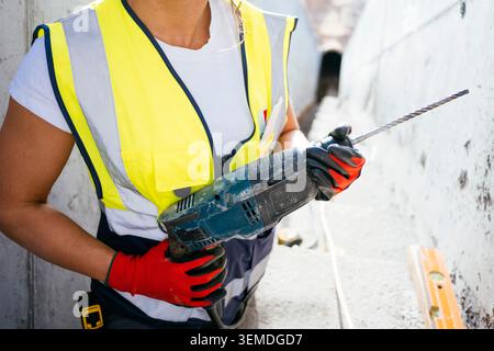 Close-up of a woman construction worker in reflective safety vest and gloves holding a rotary hammer drill with long bit at a job site, focused on ren Stock Photo