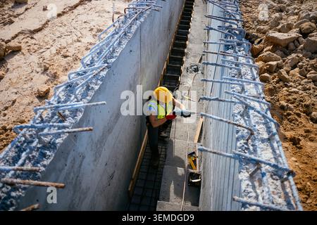 Top view of a female construction worker in hard hat and safety vest checking measurements in a concrete foundation trench with rebar, tools, and soil Stock Photo