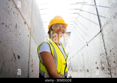 Smiling female construction worker wearing hard hat, safety glasses, and high visibility vest in a concrete tunnel, representing industry, safety, and Stock Photo