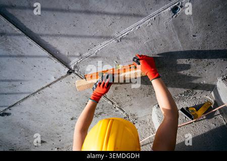 Top view of a female construction worker wearing hard hat and safety gloves, using a spirit level to align and check a rough concrete ceiling during r Stock Photo