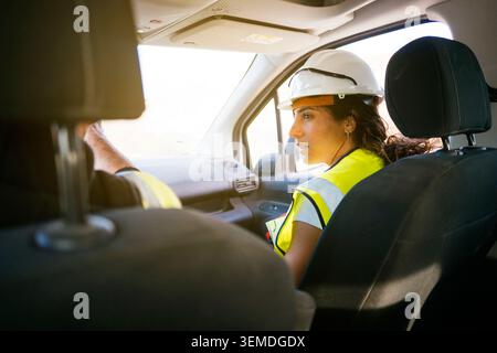 Female construction worker in reflective safety vest and hard hat sits in a vehicle, checking a clipboard while traveling to a job site during dayligh Stock Photo