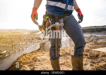 Female construction worker stands on a dusty building site at sunrise, wearing reflective vest, gloves, and tool belt with hammer and tape measure, re Stock Photo