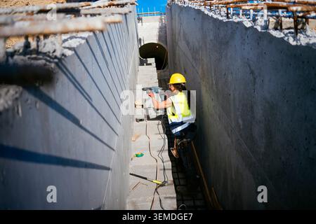Female construction worker in hard hat and safety vest uses a power drill in a concrete trench, preparing reinforcement for an underground passage on Stock Photo