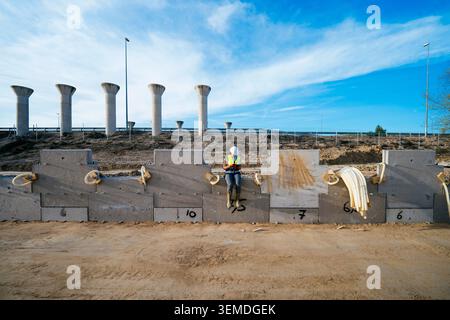 Female construction worker in hard hat and safety vest sits on a concrete barrier at an infrastructure site, with pylons, pipes, and blue sky in the b Stock Photo