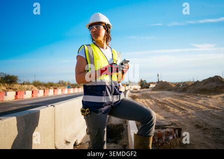 Female construction worker in hard hat and reflective vest stands by highway barriers, checking phone and surveying roadworks at a sunny outdoor const Stock Photo