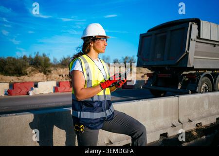 Female construction worker in hard hat and reflective vest uses a smartphone at a roadside work zone, checking project updates while a truck passes ne Stock Photo