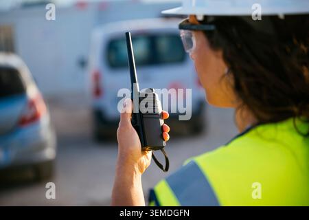 Rear view of a woman construction worker in a safety helmet and reflective vest using a two-way radio at an active work site, coordinating operations Stock Photo