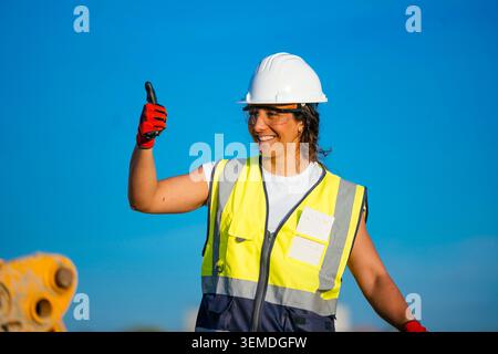 Smiling female construction worker wearing white hard hat, high visibility vest and gloves, giving thumbs up at a worksite, promoting safety, teamwork Stock Photo