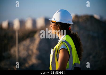 Side profile of a female construction worker wearing a hard hat, safety glasses, and reflective vest, standing at an outdoor worksite with blurred mac Stock Photo