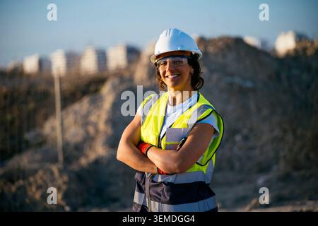 Confident female construction worker wearing a hard hat, safety glasses, and reflective vest, standing with arms crossed at an active earthworks site Stock Photo