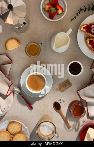 Top view of a continental breakfast table with espresso, milk, moka pot, toast with jam, biscuits, strawberries, and honey on a linen surface for a co Stock Photo