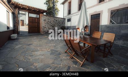 Wide-angle view of an empty stone courtyard with wooden outdoor table, folding chairs, closed umbrella, rustic wall and open wooden gate Stock Photo