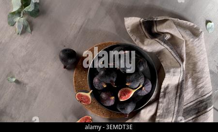 Top view of a moody food composition featuring fresh purple figs in a black bowl placed on a cork coaster. Sliced open fruit reveals a vibrant red cor Stock Photo