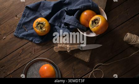 A top down view captures bright orange persimmons arranged on a dark wooden surface with a blue cloth and kitchen knife. Moody low key lighting emphas Stock Photo