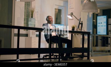 Relaxed accountant listening to music while working nightshift in office. Jolly worker entertaining colleague, making him laugh by doing dance moves and having fun at work Stock Photo