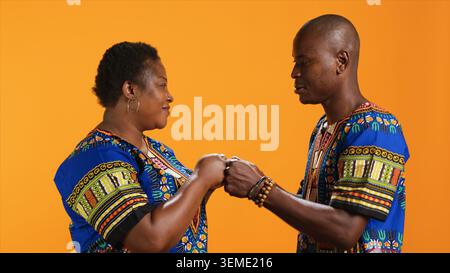 Playful ethnic couple doing fist bump gesture on camera, greeting each other with a cool modern sign in studio. Traditional african american people in colorful attire having fun over background. Stock Photo