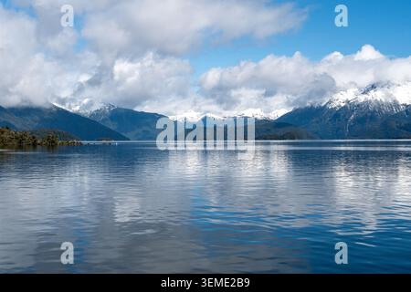 Scenic panoramic view of large, tranquil Lake Manapouri with beautiful water reflections and dramatic snow-covered mountains under a blue sky. Stock Photo