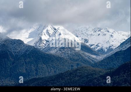 Majestic close-up view of snow-covered Remarkables mountain peaks rising above dense green forested ridges under a moody, overcast sky. Stock Photo