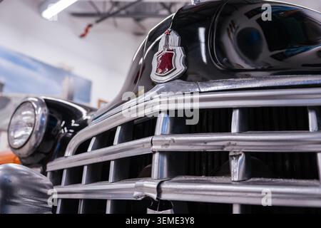 Close-up view captures front grille and hood emblem of vintage black car. Chrome grille features horizontal bars and red shield emblem, reflecting bri Stock Photo