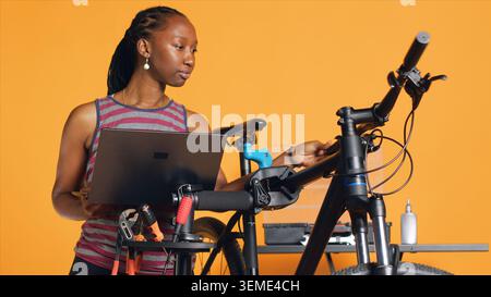 Smiling repairman doing bike servicing in studio background repair shop, looking on laptop maintenance list. Professional checking on internet for replacement bicycle parts, camera A Stock Photo
