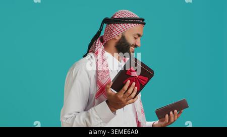Arab person opening giftbox with ribbon, receiving beautiful present on his birthday. Middle eastern happy guy feeling excited about gift wrapped with box, holiday package. Stock Photo