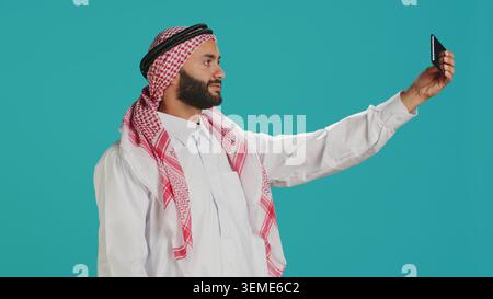 Young arab person takes photos with phone, capturing funny moments on smartphone camera in studio. Middle eastern guy with national headscarf taking pictures in traditional attire. Stock Photo
