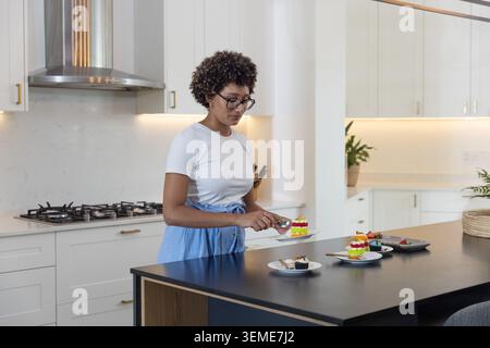Adult African American woman wearing apron plating layered cakes on kitchen island, using spoon Stock Photo