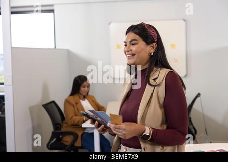 Female colleagues presenting, standing woman in maroon top holding smartphone and note at office Stock Photo