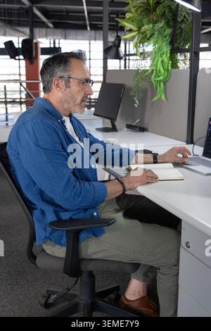 Middle-aged blue-shirted man typing on laptop at desk in open-plan office with notebook and coffee Stock Photo