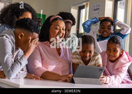 Teacher with children gathering around tablet on classroom table, wearing pink blouse and headband Stock Photo
