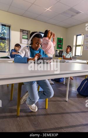 Diverse students with female teacher gesturing in classroom while boy wearing headset using tablet Stock Photo
