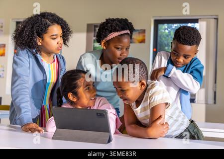 Diverse children gathering around tablet in folio on white classroom table, leaning forward Stock Photo