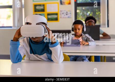 African American children sitting at desks in classroom wearing VR headset, using tablet and laptop. Pupils, learners, edtech, immersive, technology, Stock Photo