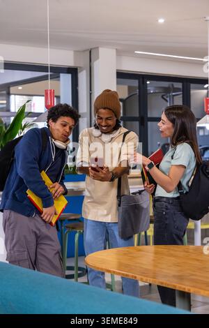 Diverse students leaning in, showing red phone in campus lounge, holding notebooks and backpacks Stock Photo